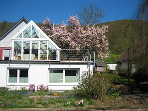 A modern house with a large glass balcony, surrounded by blooming trees and a garden. - Ferienwohnung in Lowensen mit Eigener Terrasse (Bad Pyrmont)