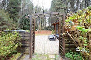 Un arc en bois mène à un chalet doté d’une terrasse en briques et d’un banc.