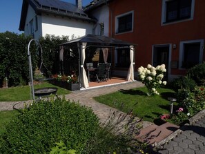 A covered outdoor seating area with a table and chairs, surrounded by a garden with various plants and flowers. - Modern Apartment in Eifel Near Forest (Dohm-Lammersdorf)