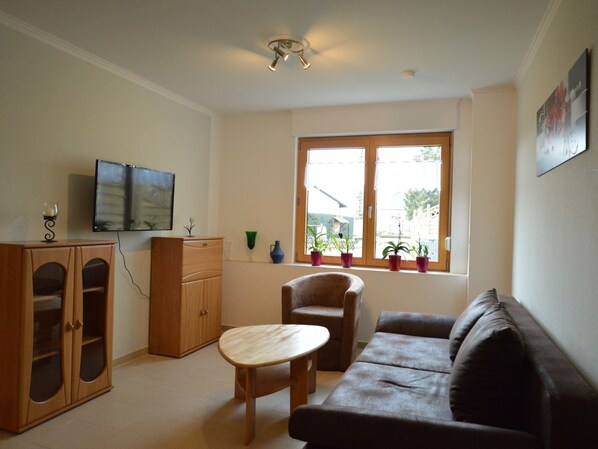 A living room with a sofa, a wooden cabinet, a small table, and a TV mounted on the wall. There is a window with potted plants on the windowsill. - Modern Apartment in Eifel Near Forest (Dohm-Lammersdorf)
