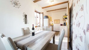 Uma área de refeições com uma mesa de madeira, cadeiras e uma garrafa de vinho. Cozinha ao fundo, com uma janela e uma porta.