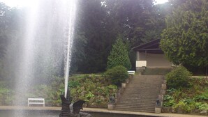 Uma fonte com estátua de dragão em um lago, rodeada por vegetação e um edifício com escadas.