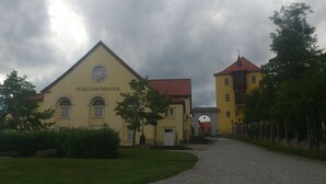 Um edifício com a inscrição 'SCHLOSSTHEATER', com um caminho de pedras que leva até ele, cercado por vegetação e sob um céu nublado.