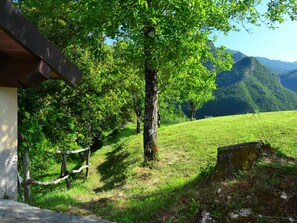 A rustic cabin with a wooden deck and a stone wall, surrounded by lush greenery and a mountain backdrop. - Belvilla by OYO Boccabugia del Colle (Fabbriche di Vergemoli)