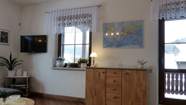 Ein Wohnzimmer mit Holzschrank, einem an der Wand montierten Fernseher, einer Globusweltkarte und einem Fenster mit Spitzenvorhängen.