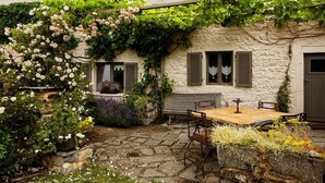 A stone patio with a wooden table and chairs, surrounded by lush greenery and flowering plants. - Charming Cottage in a Farm With Views (Doische)