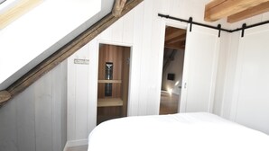 A bedroom with a sloped ceiling, a wooden shelf, a bed, and a door leading to another room. - Apartment in North Holland Near Beach (Callantsoog)