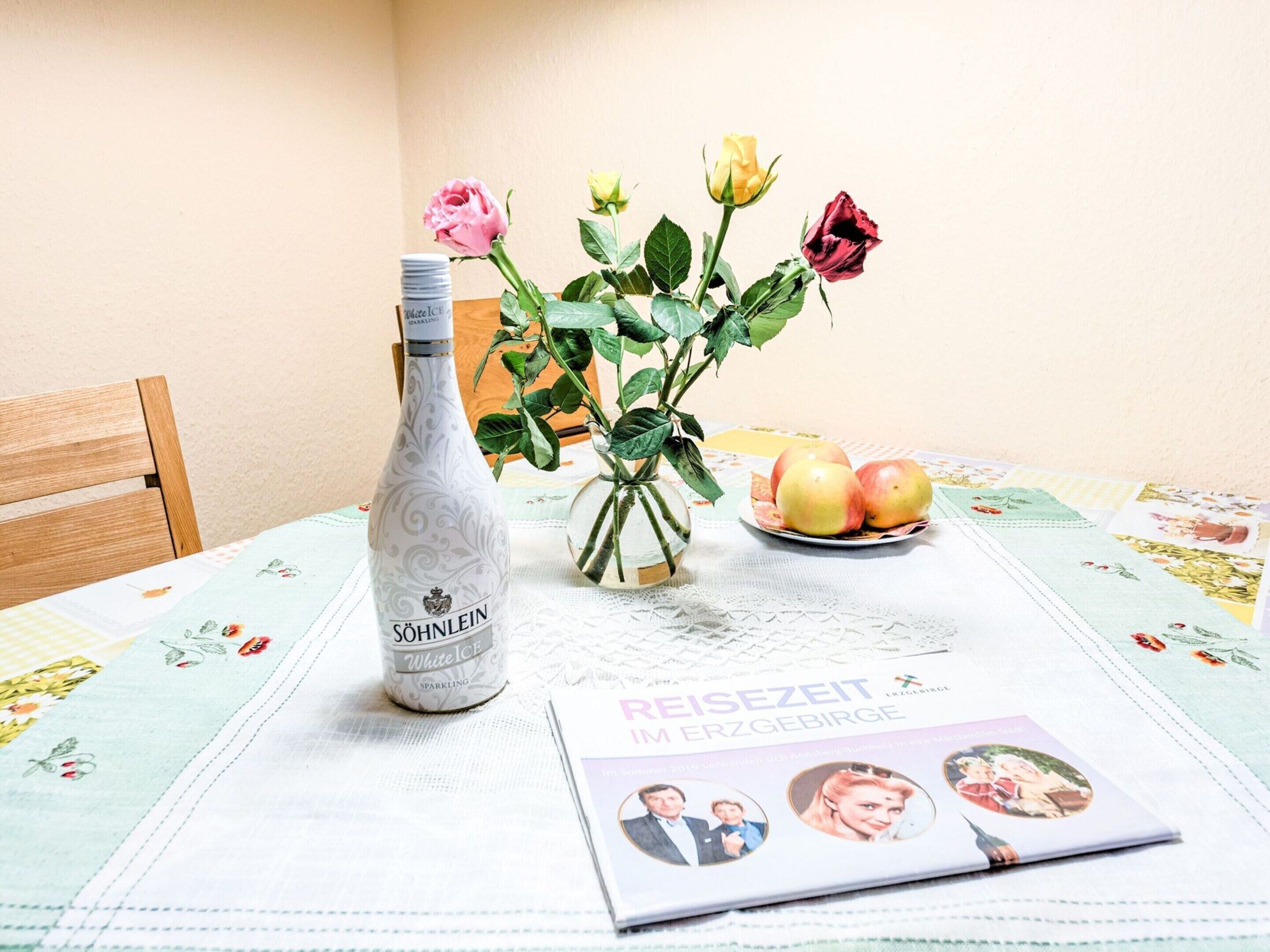 A bottle of SÖHNLEIN White Ice sparkling wine, a vase with roses and apples, and a magazine on a tablecloth with a floral pattern.