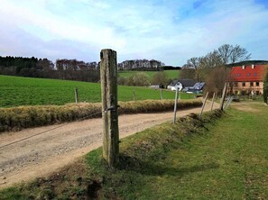 Une route de campagne bordée d’un poteau en bois et d’une clôture en fil de fer, menant à une maison équipée de panneaux solaires.