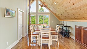 A dining area with a wooden table and chairs, a bar with stools, and a view of greenery through large windows. - SMITH TREEHOUSE LAKE LURE: CLOSE TO ASHEVILLE AND TYRON WITH KILLER VIEWS (Mill Spring)
