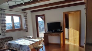 A cozy room with a wooden ceiling, a dining table with floral tablecloth, two chairs, a television on a wooden stand, and a balcony visible through a window.