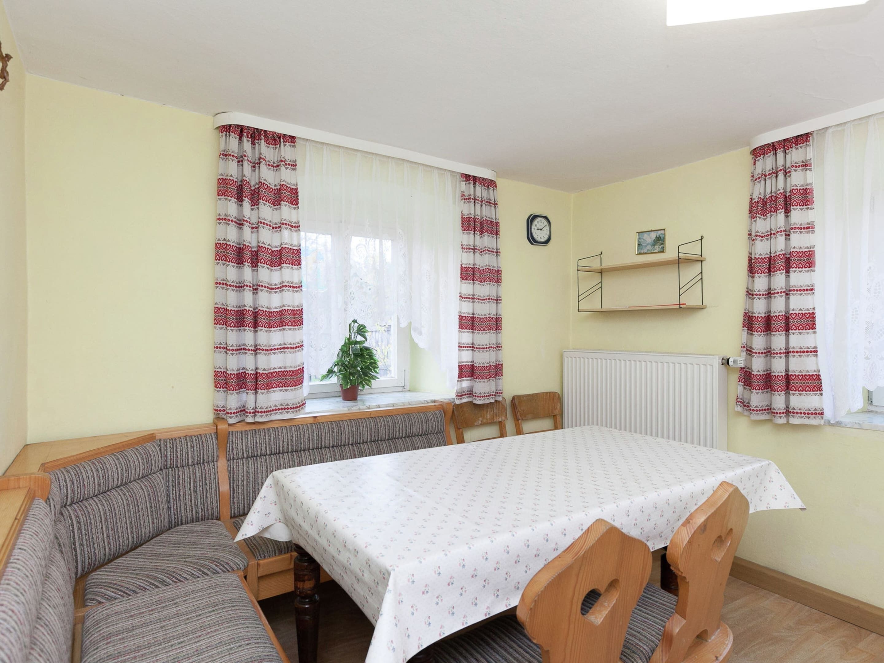A room with a dining table, two wooden chairs, a bench, a potted plant, a clock, and a radiator.