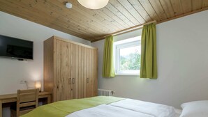 Une chambre avec un plafond en bois, un lit recouvert de draps blancs, une armoire en bois, un bureau avec une lampe et un téléviseur fixé au mur.