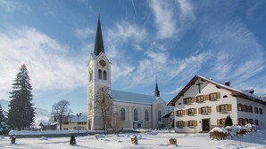 A snow-covered church with a tall steeple and a building with wooden shutters. - Apartment in Riezlern Near Ski Slope (Mittelberg)