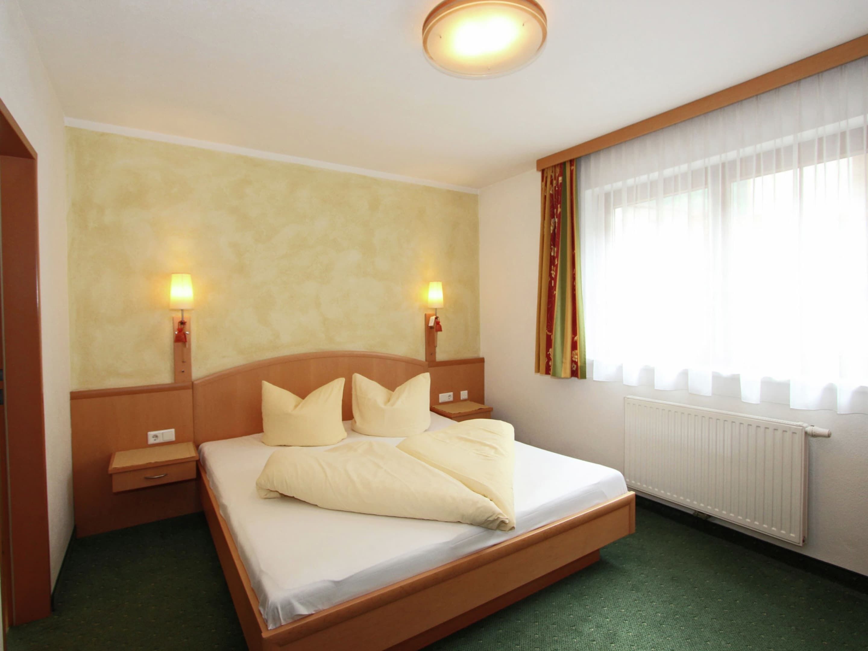 A hotel room with a single bed, two bedside tables, two wall-mounted lamps, a window with curtains, and a radiator.