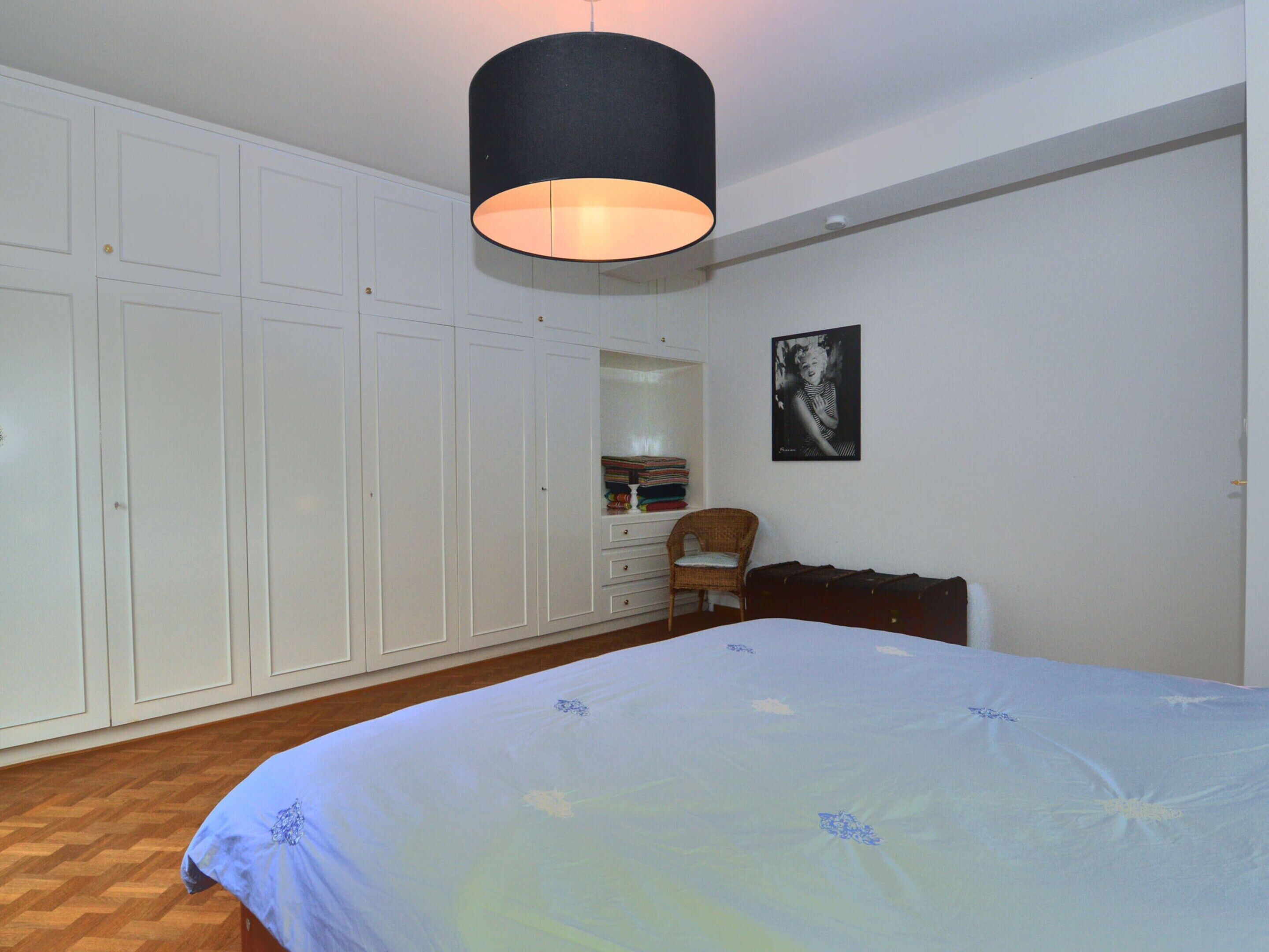 A bedroom with a large bed, a wardrobe, a chair, a desk, and a hanging lamp.