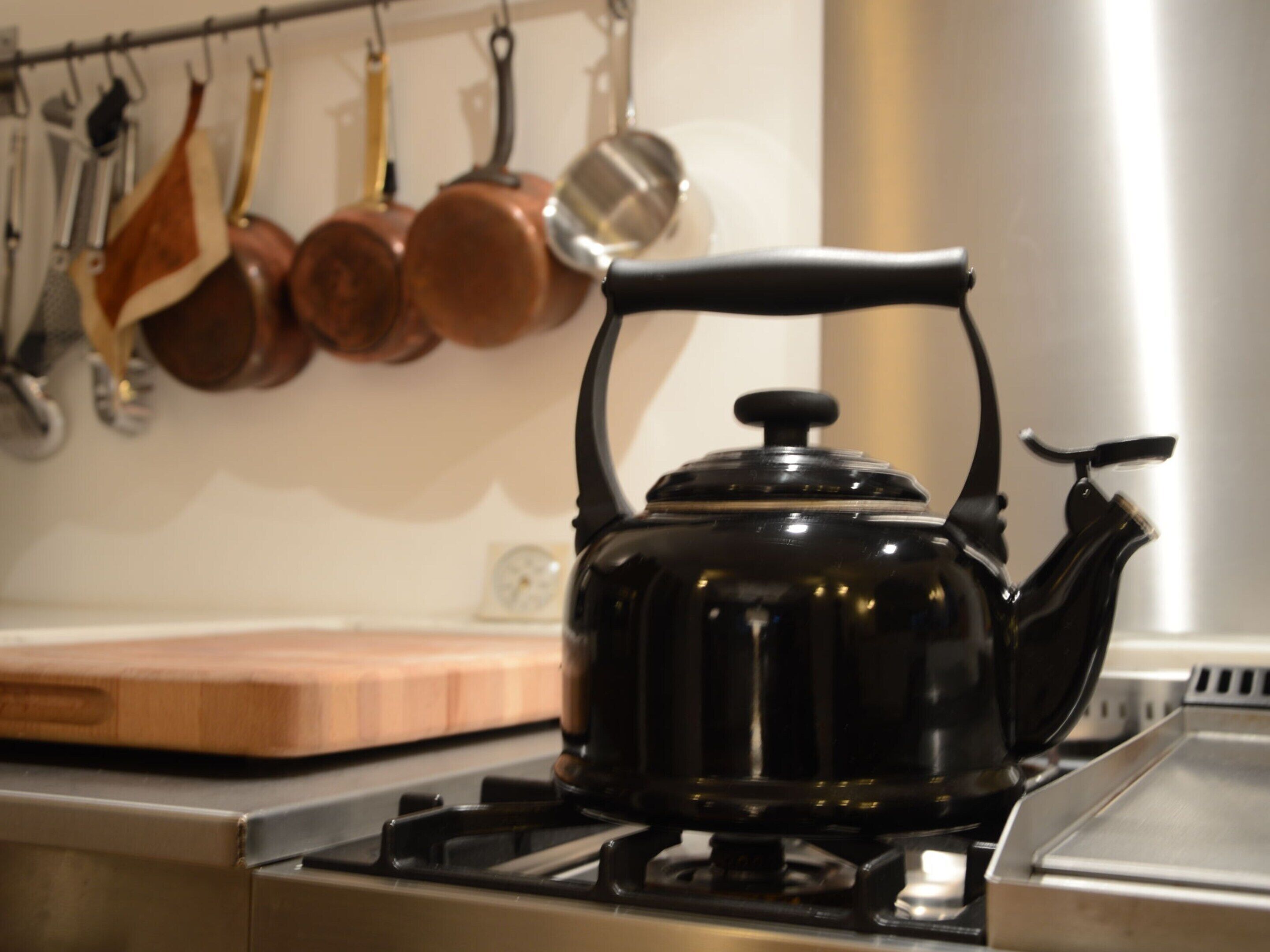 A kitchen with a black kettle on a stove, a wooden cutting board, and copper pots hanging on the wall.