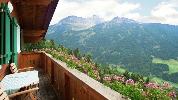 Un balcón de madera con una mesa y sillas, con vistas a un paisaje montañoso y un cielo despejado.