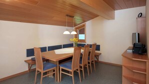 Un comedor con una mesa de madera, un banco acolchado azul y sillas. Hay una ventana con vistas, un reloj en la pared y un mueble bajo con un televisor.
