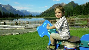 A child sitting on a blue and white horse-shaped ride in a park with a lake and mountains in the background.
