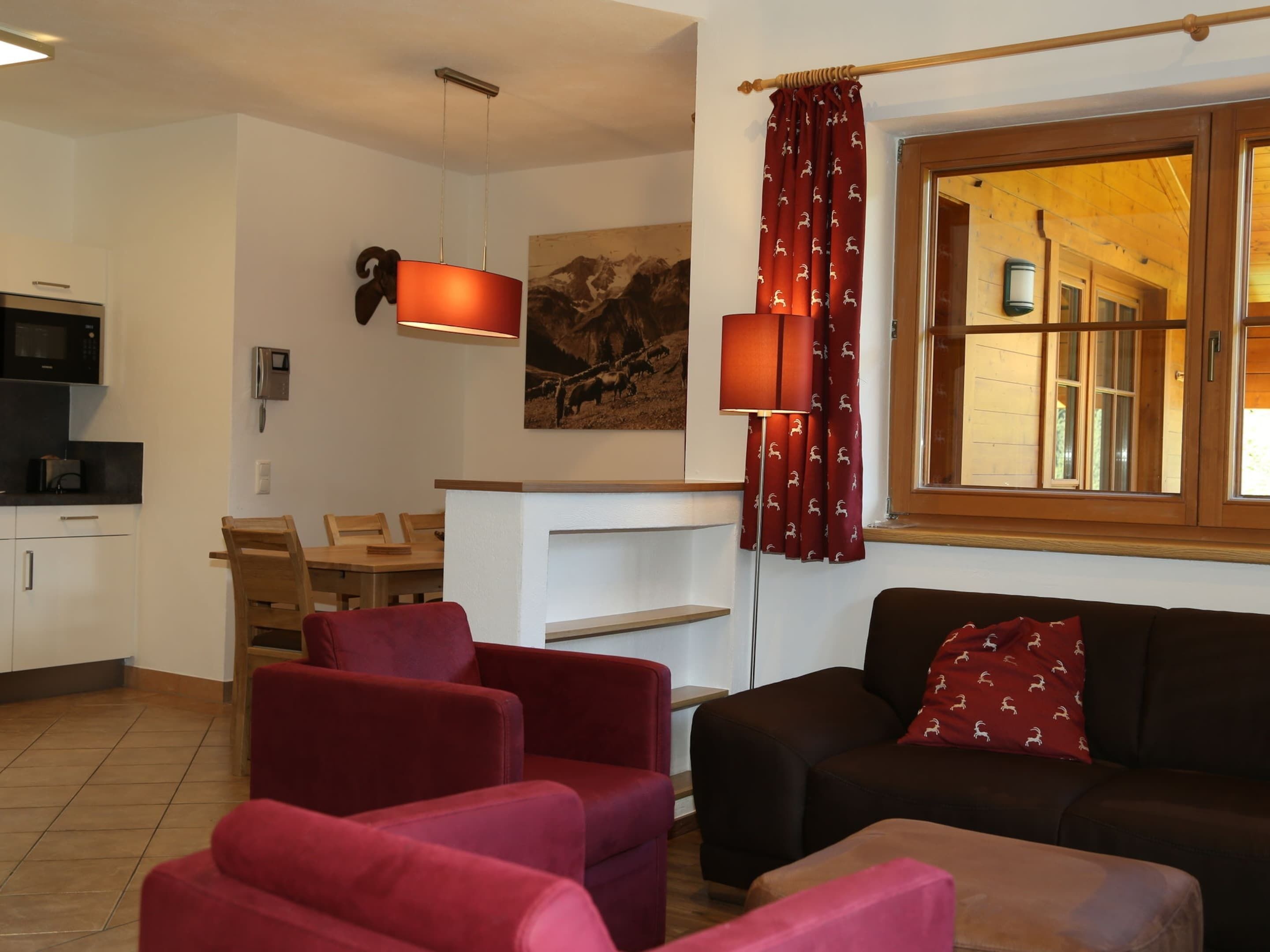 Una sala de estar con un sofá marrón, sillones rojos, una mesa de centro de madera y una cortina roja con un estampado.