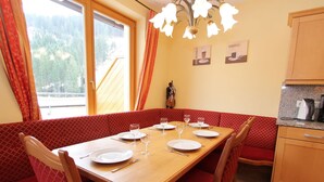 Un coin repas avec une table en bois dressée pour quatre personnes, des bancs rembourrés de coussins rouges et une vue sur l’extérieur grâce à de grandes fenêtres.