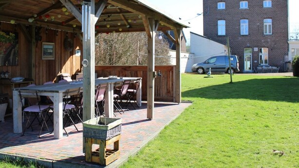 An outdoor wooden structure with a table and chairs, a covered seating area, and a brick pathway leading to a grassy area. - Spacious Holiday Home in Voeren With Spacious Garden (Voeren)