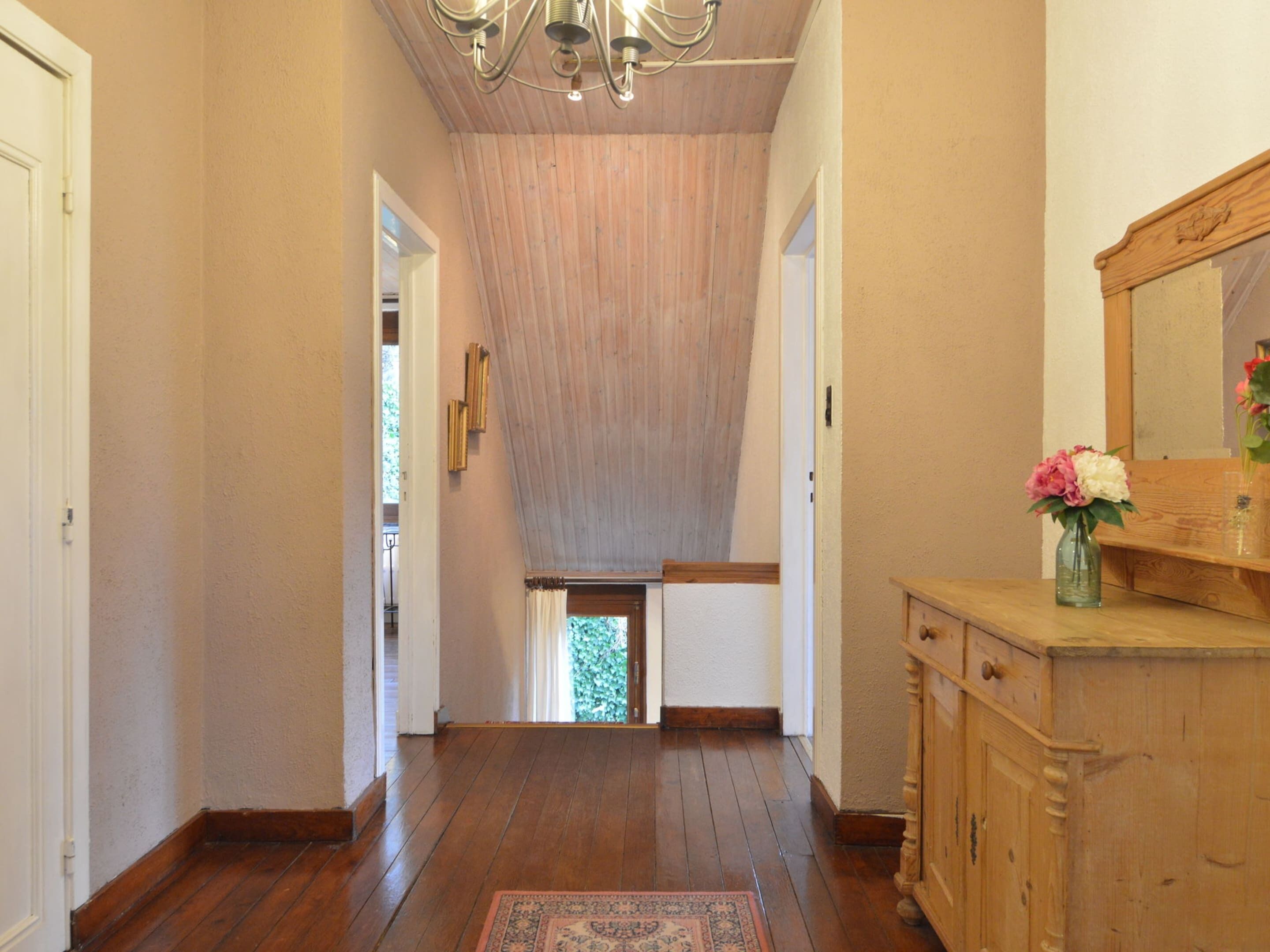 Un pasillo con suelo de madera, un candelabro, un aparador de madera con espejo, y un jarrón con flores.