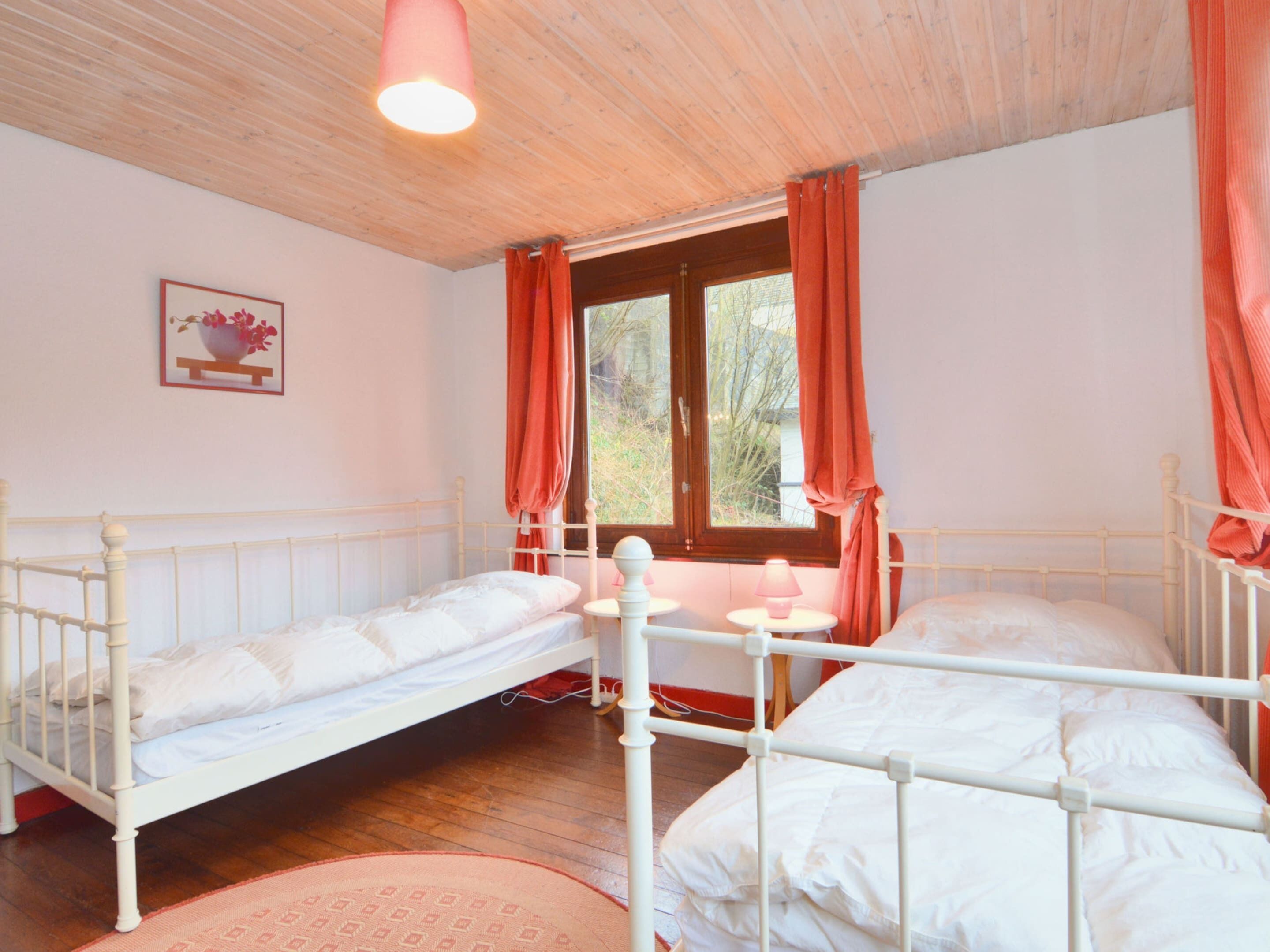 A bedroom with two single beds, a window with curtains, a small table with a lamp, and a framed picture on the wall.