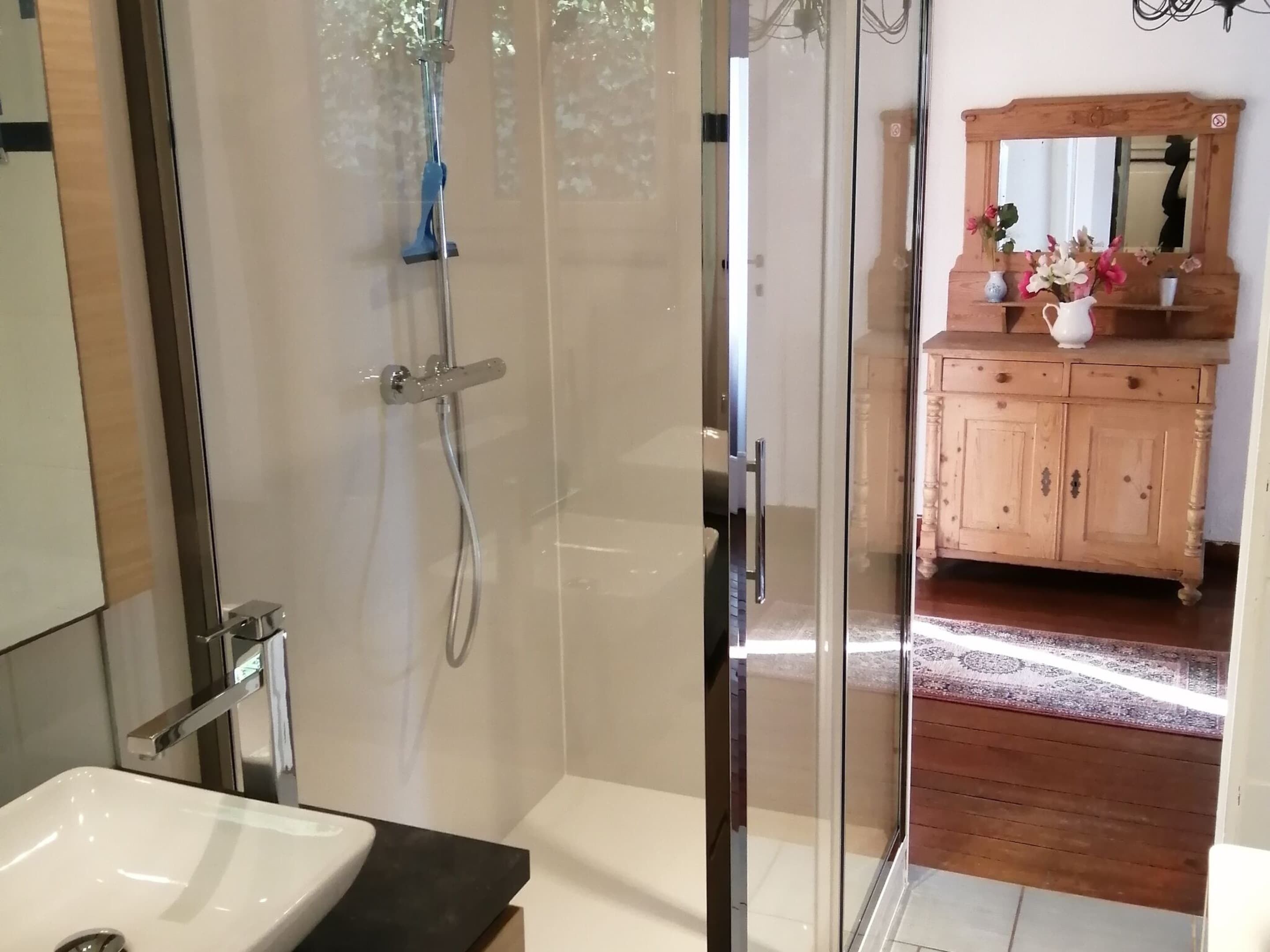 Une salle de bain moderne avec une douche à parois de verre, un lavabo blanc et un meuble-lavabo en bois agrémenté d’un miroir et d’objets décoratifs.