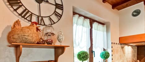 A kitchen with a large wall clock, wooden beams, and a window with curtains.