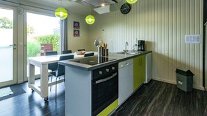 A modern kitchen with a central island, a built-in oven, and a sink. There are two pendant lights, a clock, and a small trash bin. - Boutique Chalet in Büllingen near Lake (Buellingen)