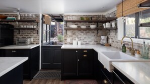 A modern kitchen with black cabinets, white countertops, and exposed brick walls. There are shelves with glassware and a coffee machine. - Palm Cottage San Diego (La Mesa)