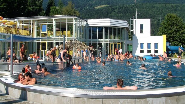 A swimming pool area with people enjoying the water, a building with large windows, and a mountainous backdrop. - Nice Apartment in Detached House with Large Garden near Town Center & Ski Slope (Kotschach-Mauthen)