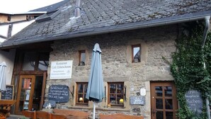 A stone building with wooden benches and tables outside, signs indicating a restaurant and coffee shop, and a small window with a view. - Cozy Holiday Home in Lesterny near Lake (Nassogne)