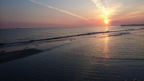 A beach scene with a sunset, ocean waves, and a pier in the distance. - Holiday Home in De Haan Near Beach (De Haan)