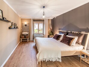 A bedroom with a wooden floor, a large bed with multiple pillows, a bedside table, a nightstand with a lamp, a window with curtains, and a wall-mounted shelf with decorative items. - Beautiful Group House With Large Garden (Gouvy)