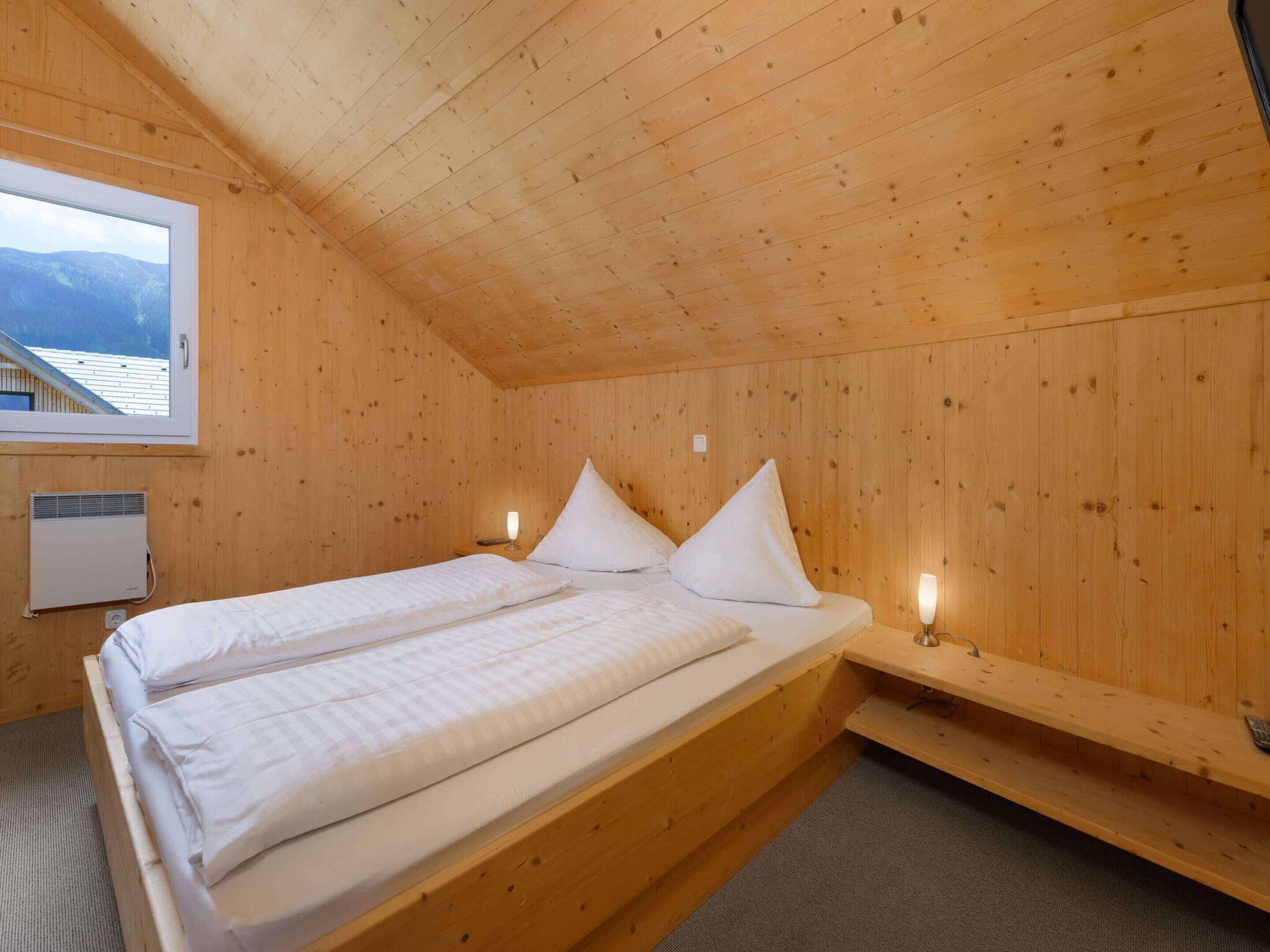 A wooden cabin room with a bed, two bedside tables, a window with a view of mountains, and a heater.