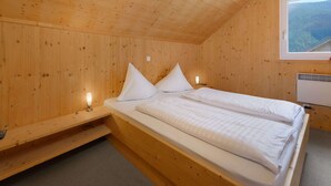 Ein Schlafzimmer einer Holzhütte mit einem Einzelbett, einem Nachttisch, einer Lampe und einem Fenster mit Blick auf einen bewaldeten Bereich.