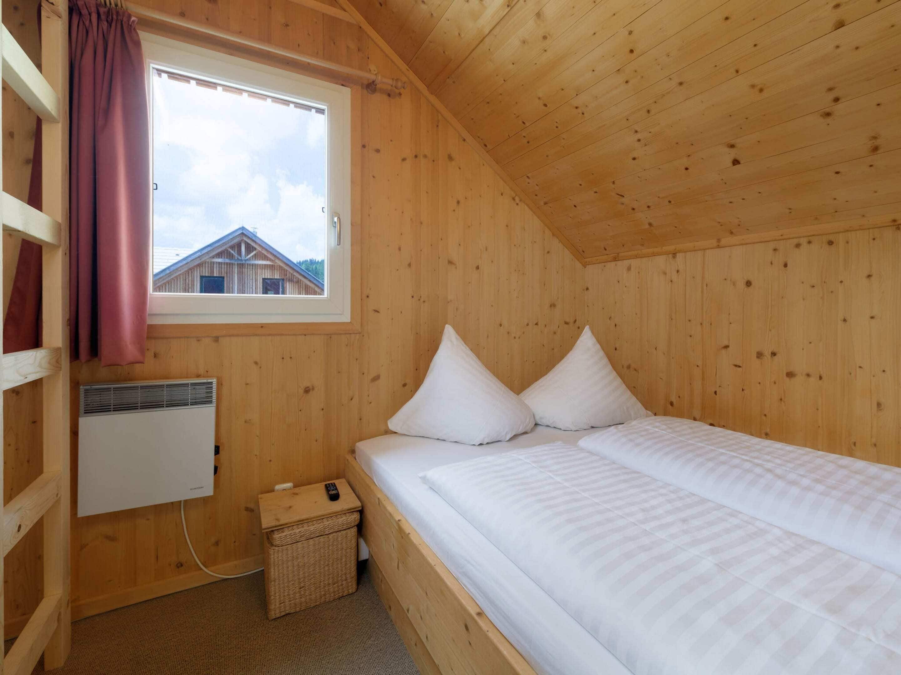 A wooden cabin room with a bed, a window, a radiator, and a small basket.