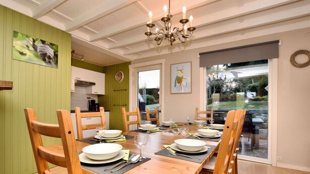 A dining area with a wooden table set for four, surrounded by wooden chairs. A chandelier hangs from the ceiling, and there is a view of a garden through a glass door. - Family Retreat With Garden (Tenneville)