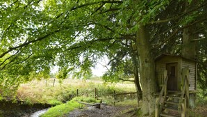 A rustic wooden structure with a ladder leading up to a door, surrounded by lush greenery and a small stream. - Elegant Beverce Escape (Malmedy)