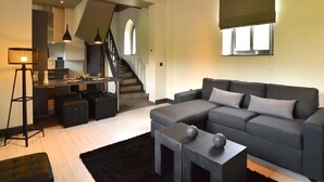 Ein modernes Wohnzimmer mit einem grauen Sofa, einem schwarzen Ottomanen und einem kleinen Beistelltisch mit dekorativen Kugeln. Eine Treppe führt zu einer oberen Ebene, auf der eine Küche erkennbar ist.