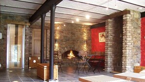 Ein Kamin aus Stein mit Feuer, hölzerne Stühle und ein Holztisch in einem Raum mit Stehwänden und Fliesenboden.