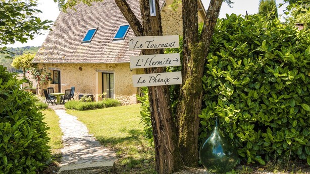 Ein Steinhaus mit Ziegeldach, ein Weg, der zu einem Tisch und Stühlen führt, und Hinweisschilder.