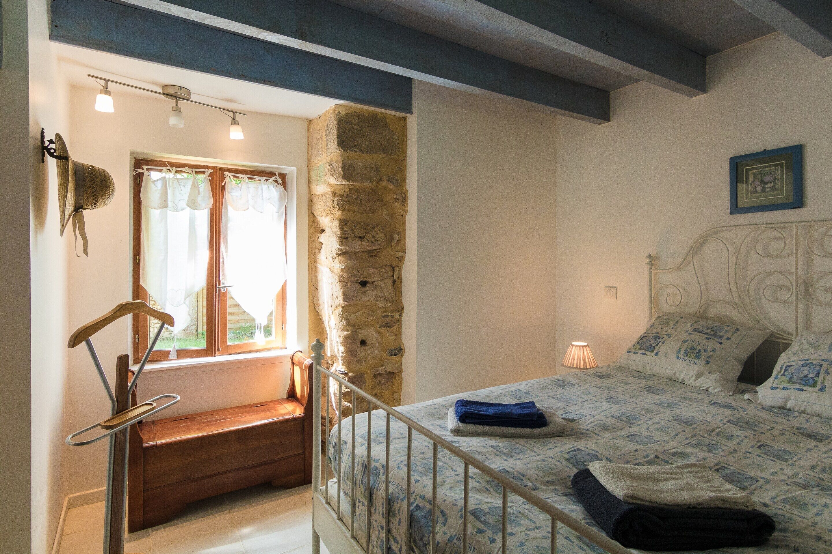 A bedroom with a bed, a bench, a stone wall, and a window with curtains.