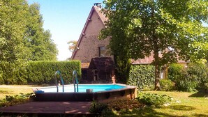 Ein Schwimmbecken mit Holzterrasse, umgeben von Grünpflanzen und einem Steinhaus mit Satteldach.