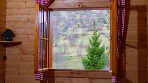 A wooden cabin room with a window featuring red and white checkered curtains, a lamp on a shelf, and a view of a forested landscape. - Drevenice Oscadnica (Oscadnica)