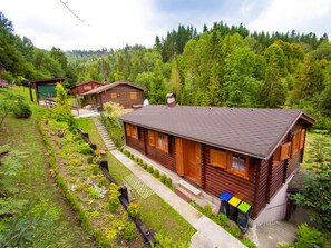 A wooden cabin with a sloped roof, surrounded by lush greenery and a forested hillside. - Drevenice Oscadnica (Oscadnica)