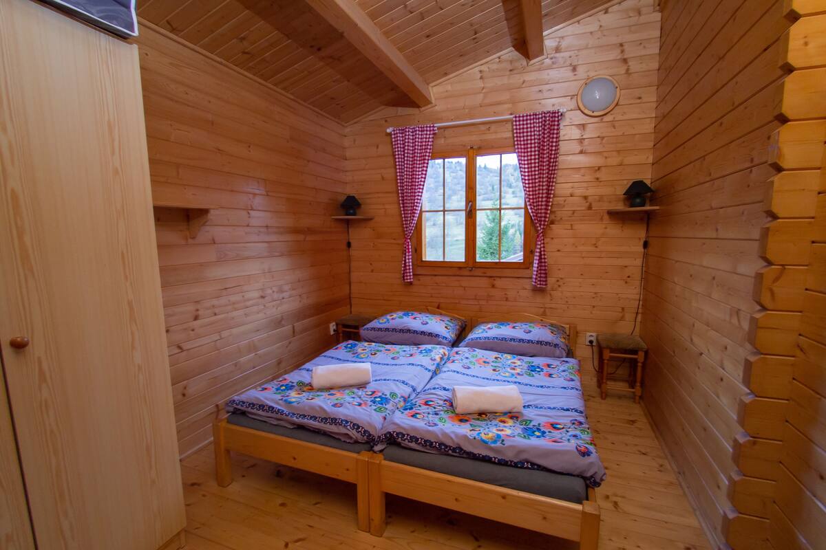 Ein Zimmer einer Holzhütte mit einem Bett, zwei Nachttischen, einem Fenster mit roten und weißen Karomustert-Vorhängen und einem kleinen Regal mit einer Lampe.